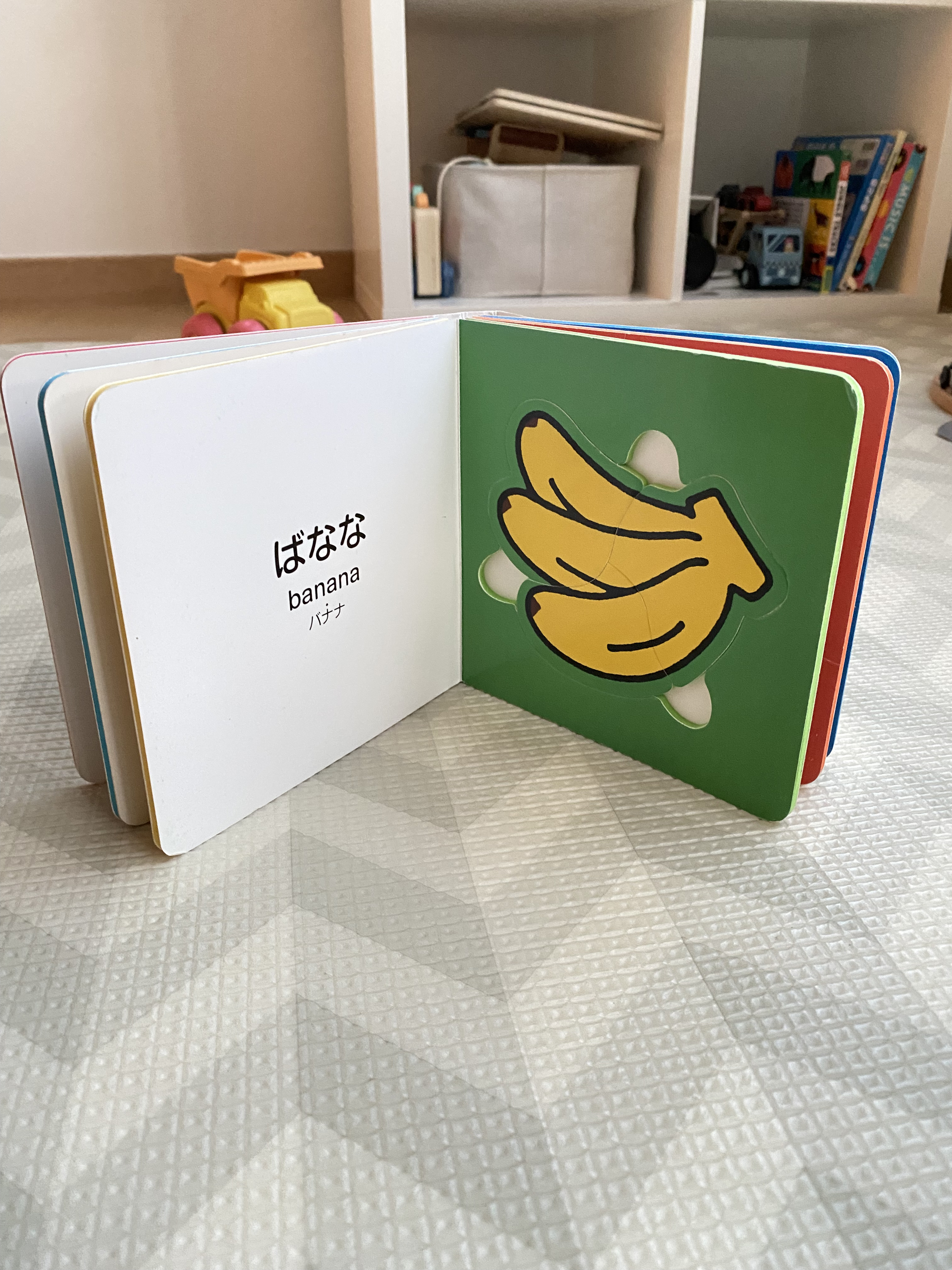 A photo of a child's book, open and standing on a mat on the floor, with a banana on the right page, and the Japanese and English word for banana on the left